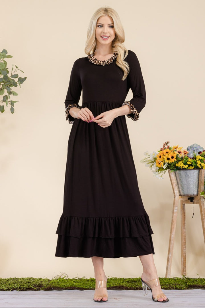 A woman stands smiling in a Celeste Full Size Leopard Ruffle Detail Midi Dress, her long blonde hair cascading down. At her side, a metal vase holds colorful flowers, and a leafy plant subtly enhances the backdrop.