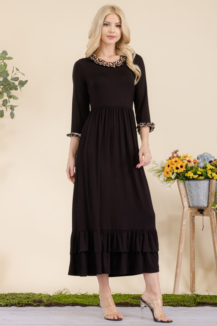 A woman with long blonde hair stands next to a wooden stool holding colorful flowers, wearing the Celeste Full Size Leopard Ruffle Detail Midi Dress, an elegant A-line black dress with leopard print accents on the collar and sleeves, and charming ruffle details at the hem.