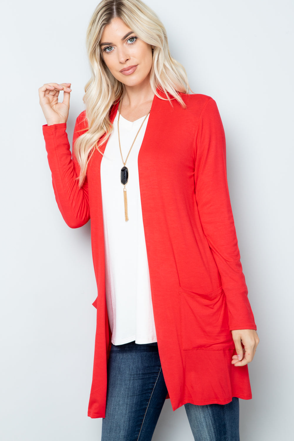 A woman with long blonde hair wears the Celeste Full Size Open Front Cardigan with Pockets in a vibrant red over a white top and jeans. She accessorizes with a long necklace featuring a black stone pendant, making her chic style stand out against the plain light-colored background.