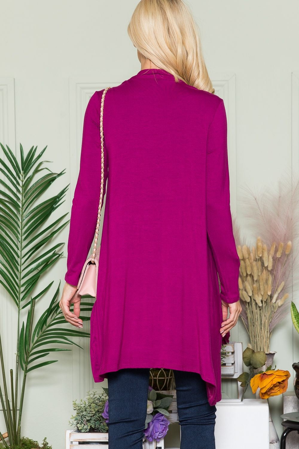 A person with long blonde hair stands with their back to the camera, wearing a vivid magenta Celeste Full Size Open Front Cardigan with Pockets. They hold a light pink purse, while green and beige plants beautifully complement the floral decor in the background.