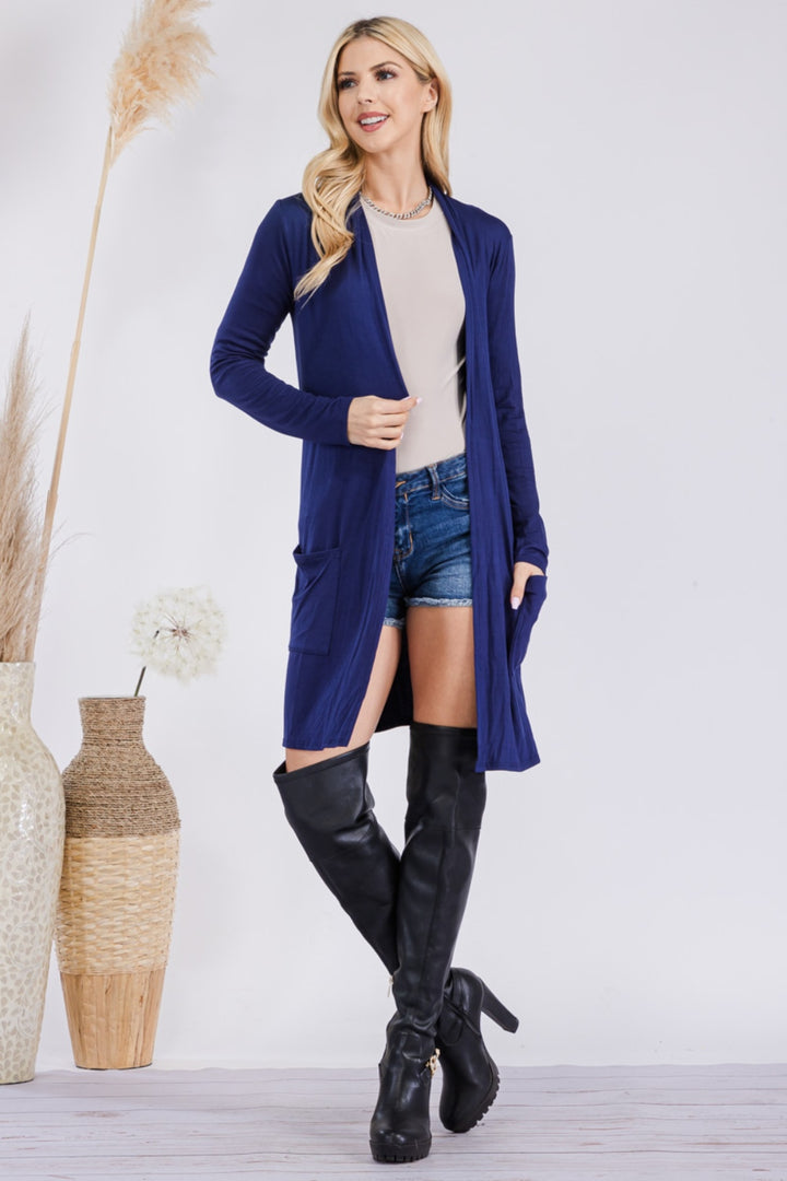 A woman confidently poses in the Celeste Full Size Open Front Cardigan with Pockets, a versatile long blue piece, layered over a beige top with denim shorts and black knee-high boots. She stands near vases of beige pampas grass against a plain background.