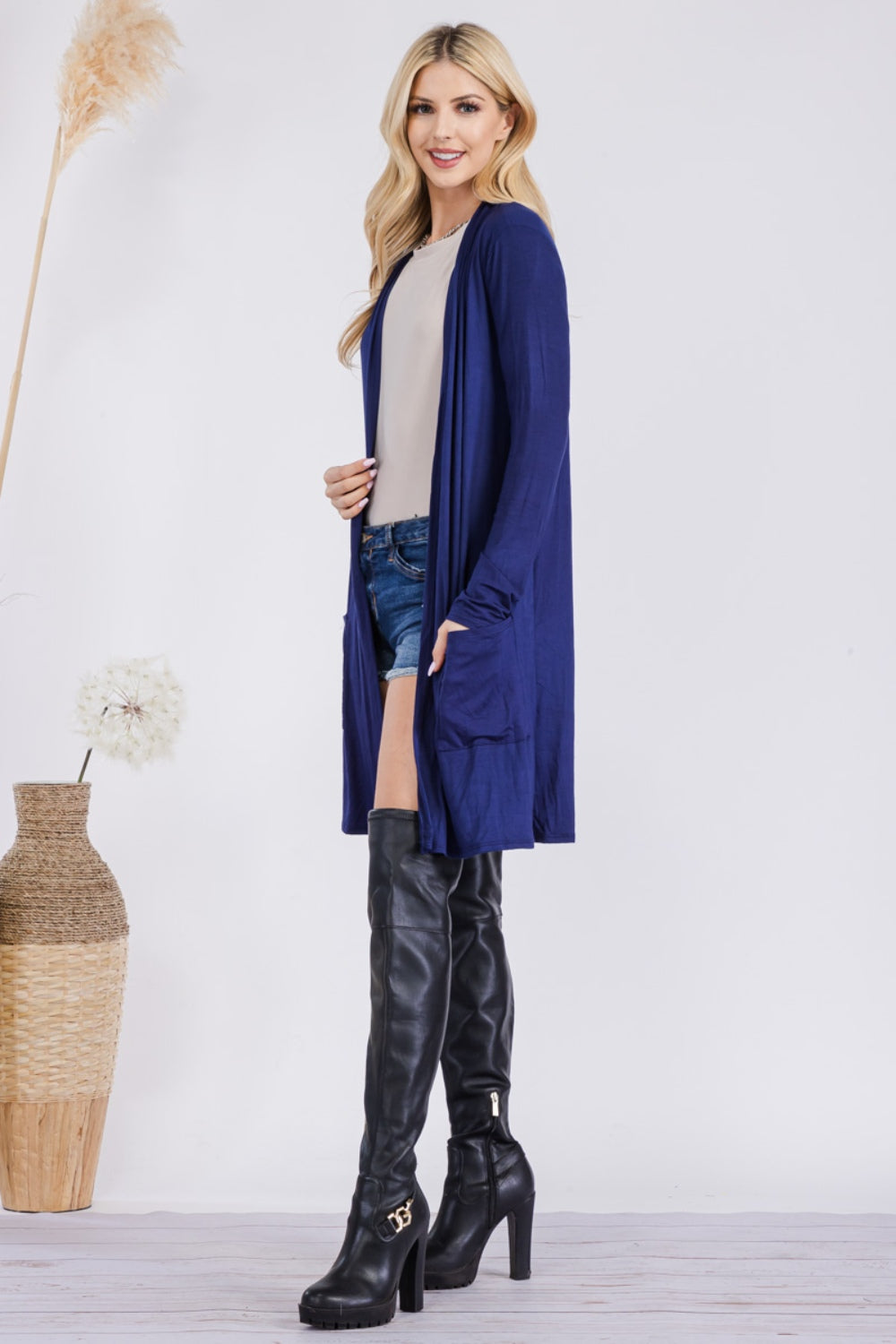 A person with long blond hair stands sideways indoors, wearing the versatile Celeste Full Size Open Front Cardigan with Pockets in blue over a light top, paired with denim shorts and black knee-high boots. Nearby are a decorative woven basket and fluffy pampas grass.