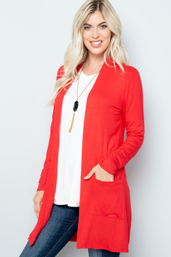 A smiling person with long blonde hair wears the Celeste Full Size Open Front Cardigan in bright red over a white top and jeans, accessorized with a long necklace. Their hand rests in a pocket against a light gray background.