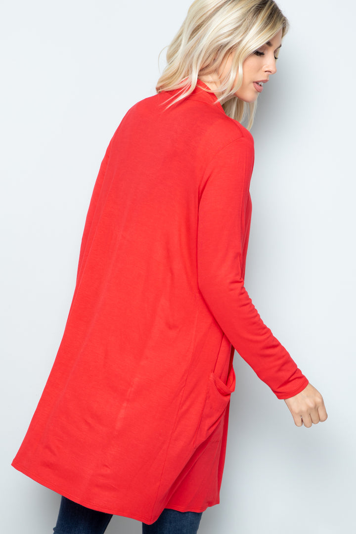 A person with blonde hair is wearing the Celeste Full Size Open Front Cardigan with Pockets in red, a versatile layering piece, over dark pants. They are turned slightly sideways, highlighting the cardigans pocket, against an off-white background with a neutral expression.