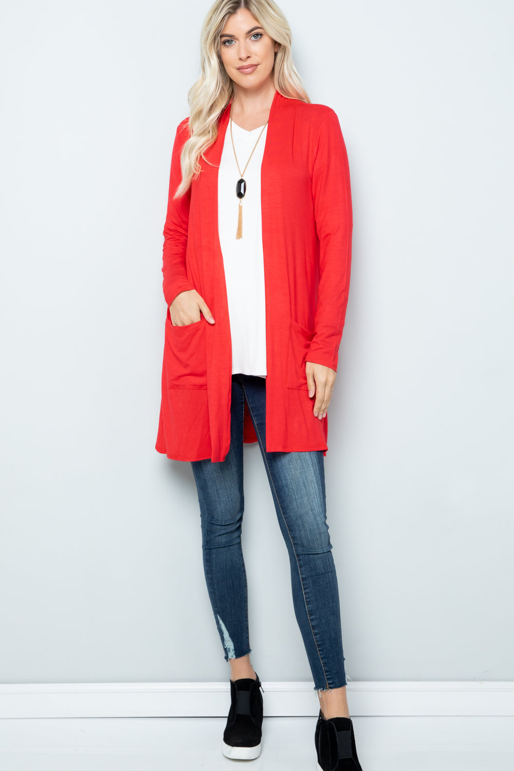 A woman with long blonde hair stands against a light grey wall, wearing the Celeste Full Size Open Front Cardigan with Pockets. This bright red versatile piece is layered over a white top, paired with blue jeans and black slip-on shoes. Her hands are casually tucked into its pockets.