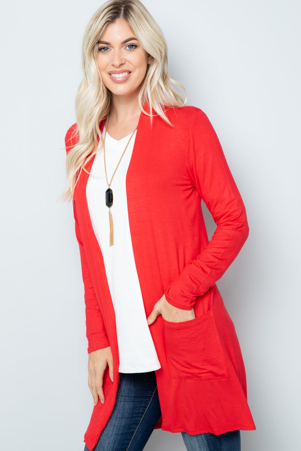 A person with long blonde hair smiles, wearing the Celeste Full Size Open Front Cardigan with Pockets in vibrant red over a white top and jeans. With a long necklace and hands in the cardigan pockets, they stand against a plain background. This versatile layering piece truly complements their style.