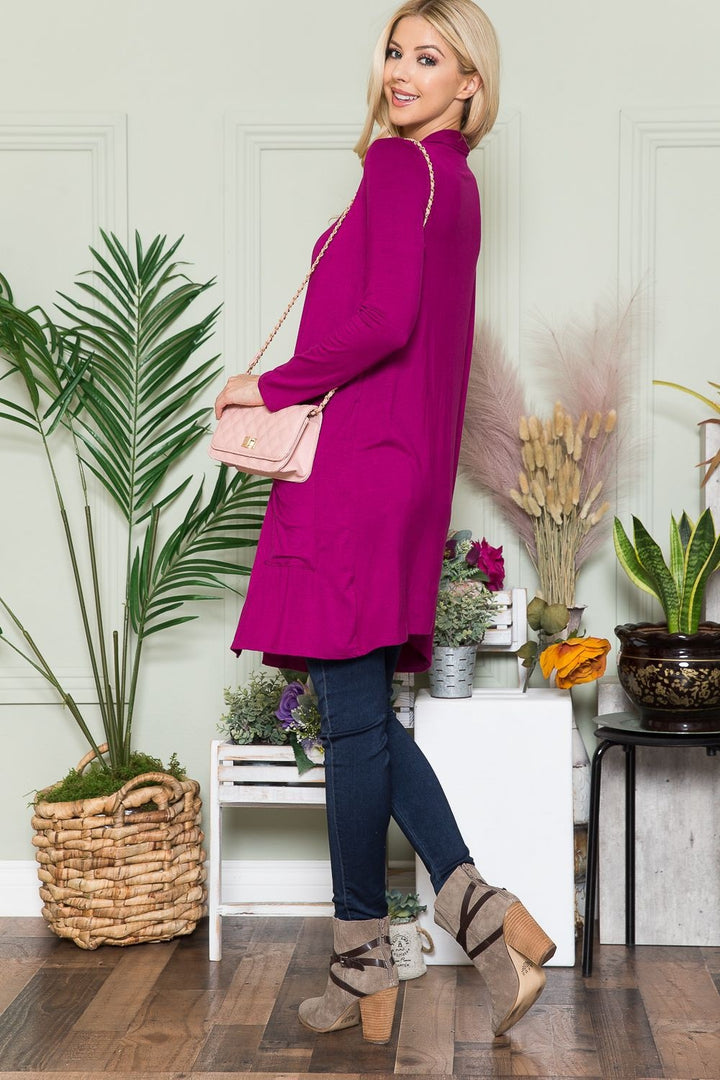 Sporting a magenta top paired with the Celeste Full Size Open Front Cardigan with Pockets, an individual poses in a room adorned with plants and decorative grass. They are wearing jeans and high-heeled shoes, holding a light pink purse, smiling, and glancing over their shoulder.