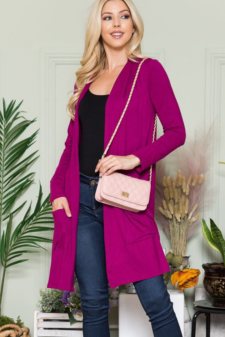 A woman wearing the Celeste Full Size Open Front Cardigan with Pockets in a vibrant pink hue over a black top and jeans stands in front of a green wall adorned with plant decor. She's holding a light pink quilted purse with a chain strap. This versatile layering piece adds both style and comfort to her look.