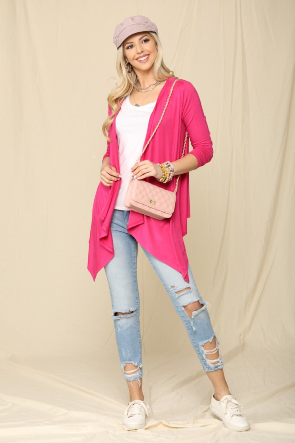 A smiling woman poses against a beige backdrop wearing the Celeste Full Size Open Front Knit Cardigan in pink, paired with a white top, ripped jeans, and white sneakers. She accessorizes with a light pink cap, matching crossbody bag, and bracelets, highlighting her versatile wardrobe staple.