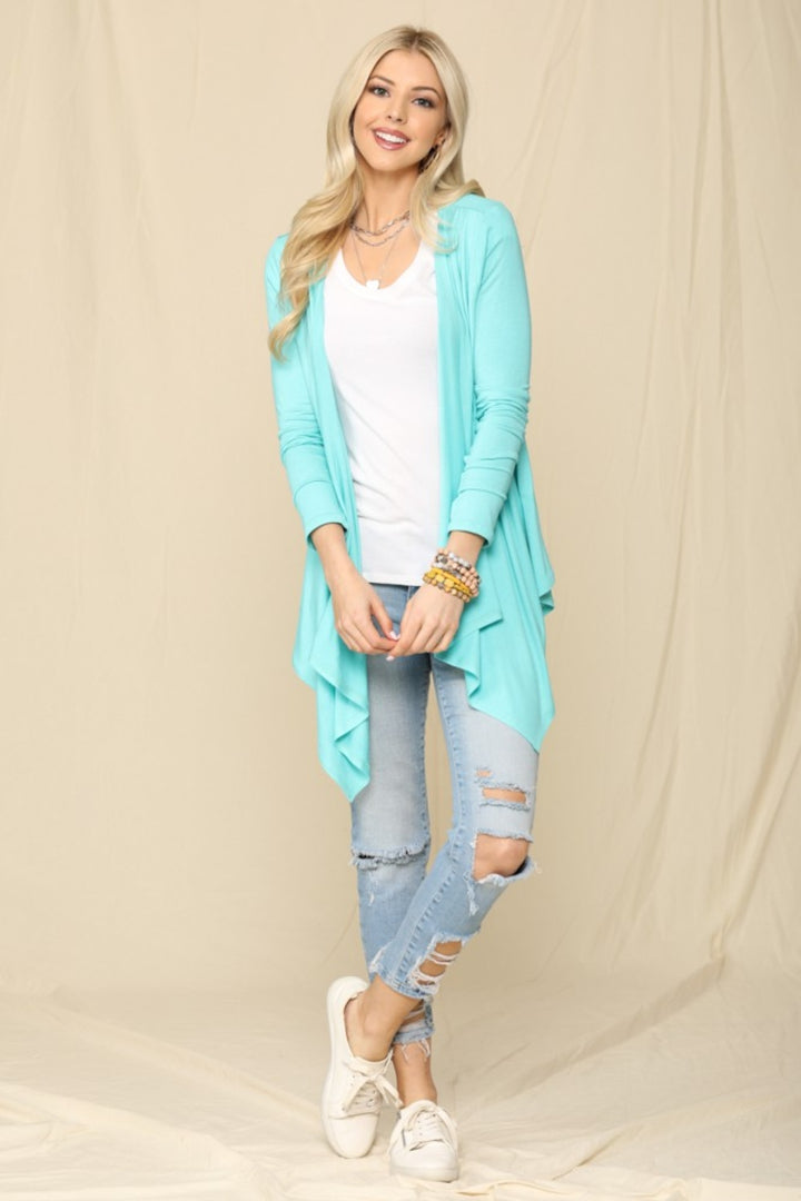 A person with long blonde hair is wearing the Celeste Full Size Open Front Knit Cardigan over a white top, distressed light blue jeans, and white sneakers, standing and smiling against a light beige backdrop.