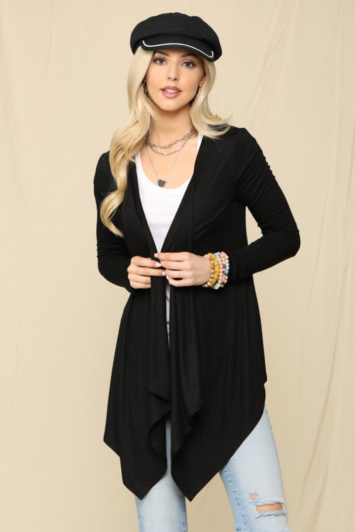 A woman in a black hat and cozy knit Celeste Full Size Open Front Cardigan over a white top stands against a beige background. Shes in light blue ripped jeans and necklaces, with her hands resting in front of her. This versatile wardrobe staple effortlessly combines style with comfort.