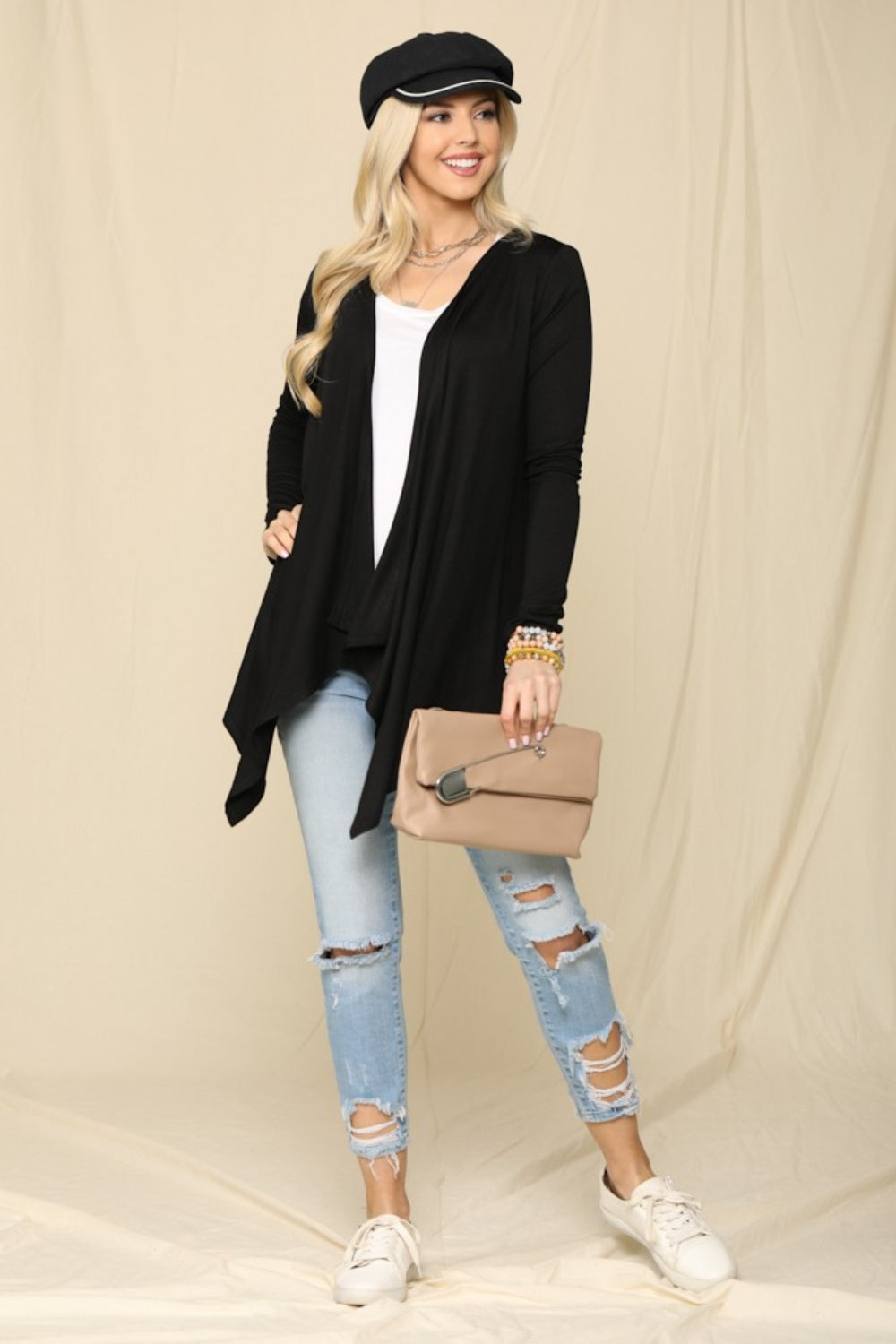 Wearing a black cap and the Celeste Full Size Open Front Knit Cardigan over a white top, a woman poses with a beige clutch. Her ripped light blue jeans add flair against a beige backdrop as she sports white sneakers and colorful bracelets, perfect for effortless style.