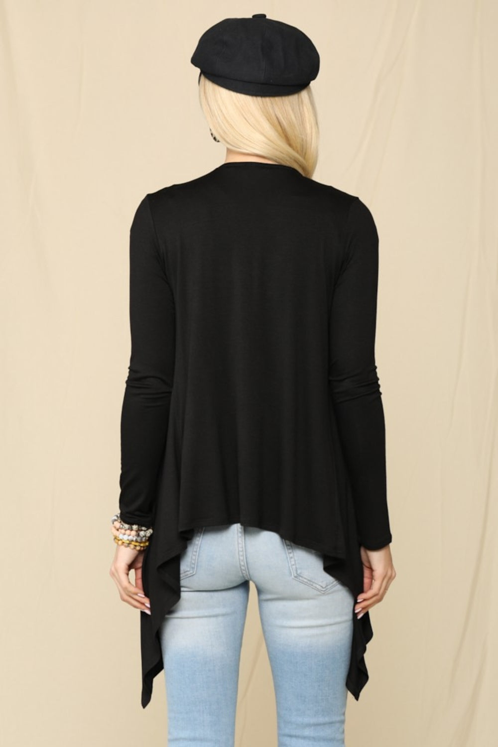 A person with long blonde hair, wearing a black beret, the Celeste Full Size Open Front Knit Cardigan in cozy black fabric, and light blue jeans is facing away. The versatile look is set against a beige background.