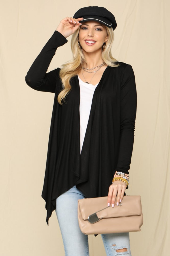 A woman in a cozy knit outfit stands against a neutral background, wearing a black hat, the Celeste Full Size Open Front Knit Cardigan, a white top, and light jeans. She carries a beige clutch and accessorizes with bracelets.