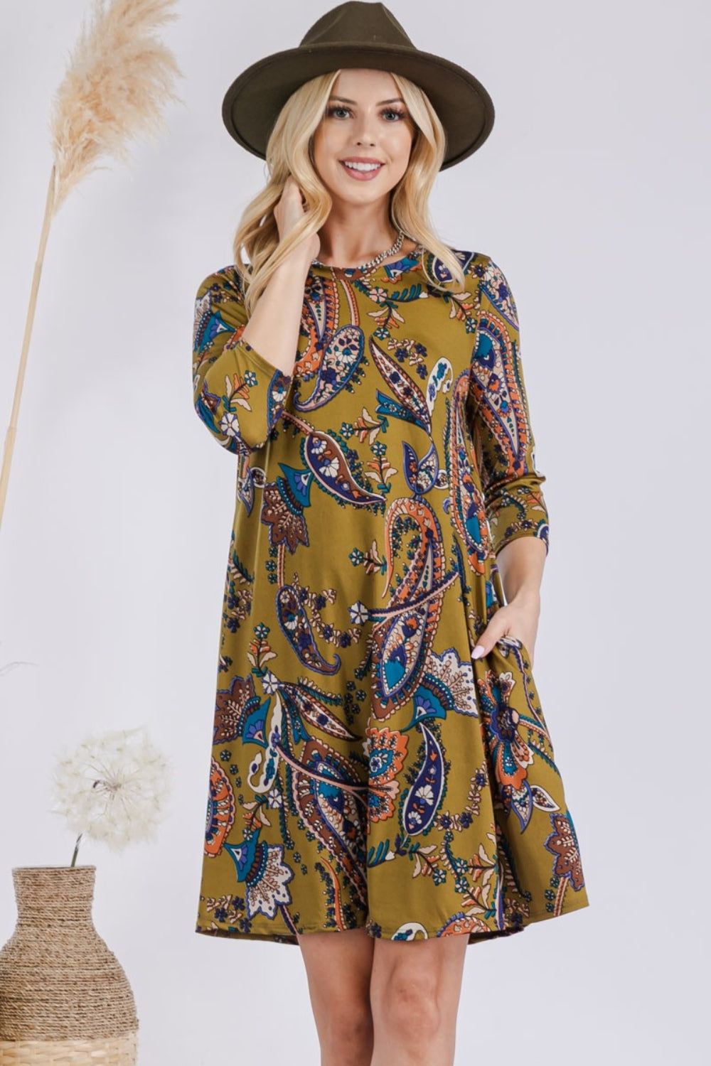 A smiling woman in a trendy Celeste Full Size Paisley Print Round Neck Dress with pockets and a wide-brimmed hat stands with one hand in her pocket next to a wicker basket decorated with dried grass and a white flower.