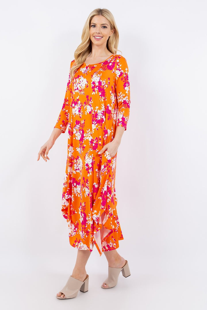 A confident woman stands against a white background in the Celeste Full Size Pick-Up Hem Asymmetric Floral Midi Dress, featuring a vibrant orange floral design, long sleeves, and an asymmetric hem. She smiles with one hand on her hip and pairs the look with light-colored open-toe sandals.