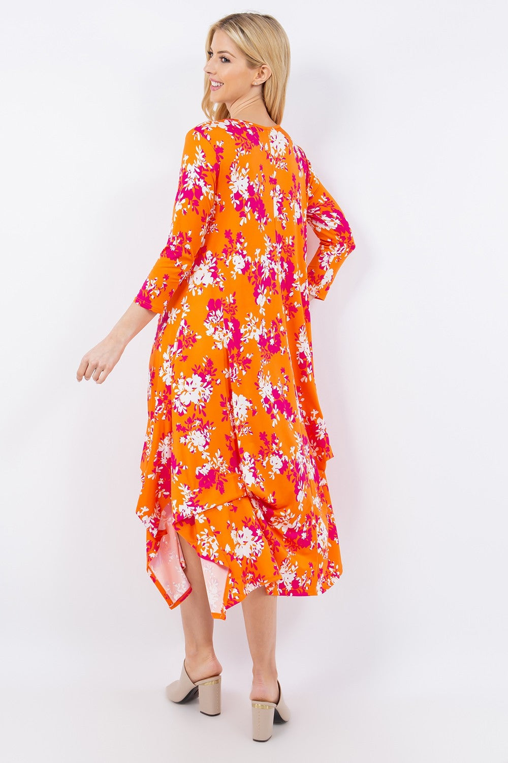 Elegantly showcasing the Celeste Full Size Pick-Up Hem Asymmetric Floral Midi Dress, a model wears vivid orange with pink and white flowers on a pure white backdrop. The dress features long sleeves and a flowy, layered style, paired perfectly with beige open-toe heels.