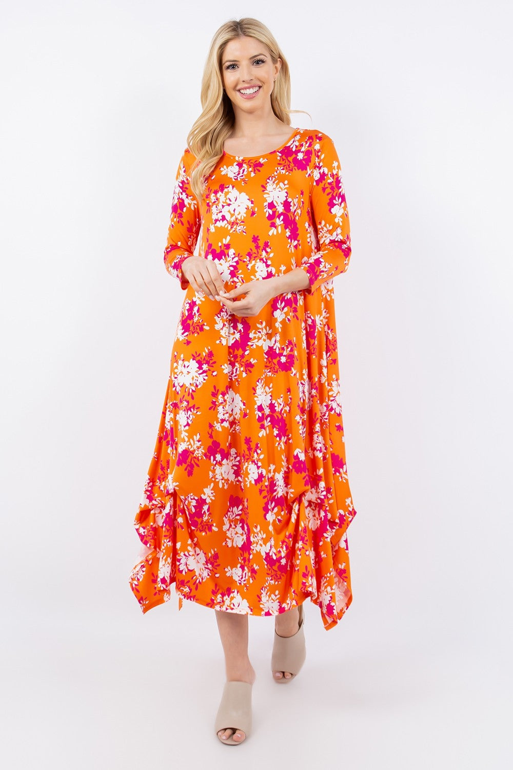 Someone is wearing the vibrant Celeste Full Size Pick-Up Hem Asymmetric Floral Midi Dress, notable for its pink and white floral pattern on orange. Styled with beige open-toed shoes, they stand against a plain white background.