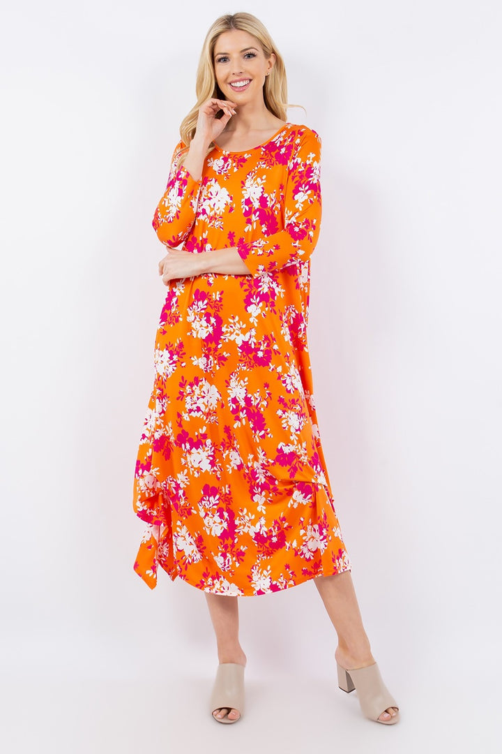 A woman models the Celeste Full Size Pick-Up Hem Asymmetric Floral Midi Dress, featuring a bright orange fabric adorned with pink and white flowers. The long-sleeved dress is elegantly paired with light-colored heeled sandals against a plain white background.