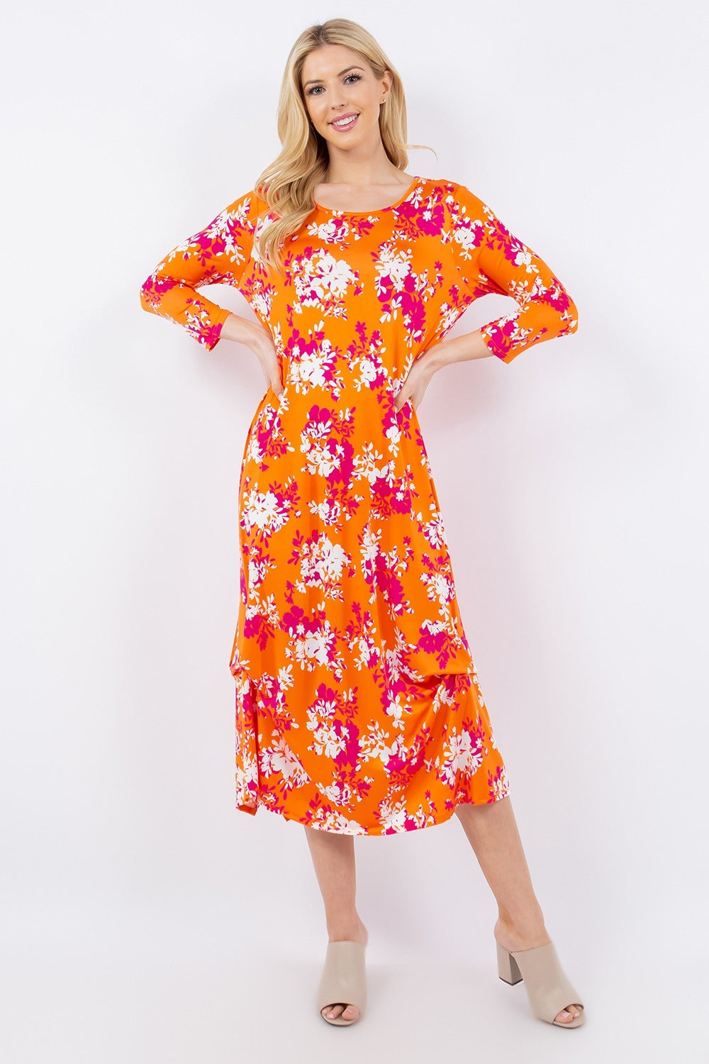 A person models the Celeste Full Size Pick-Up Hem Asymmetric Floral Midi Dress in vibrant orange with pink and white florals, posing confidently with hands on hips. Beige heeled mules complement the look against a plain white background.