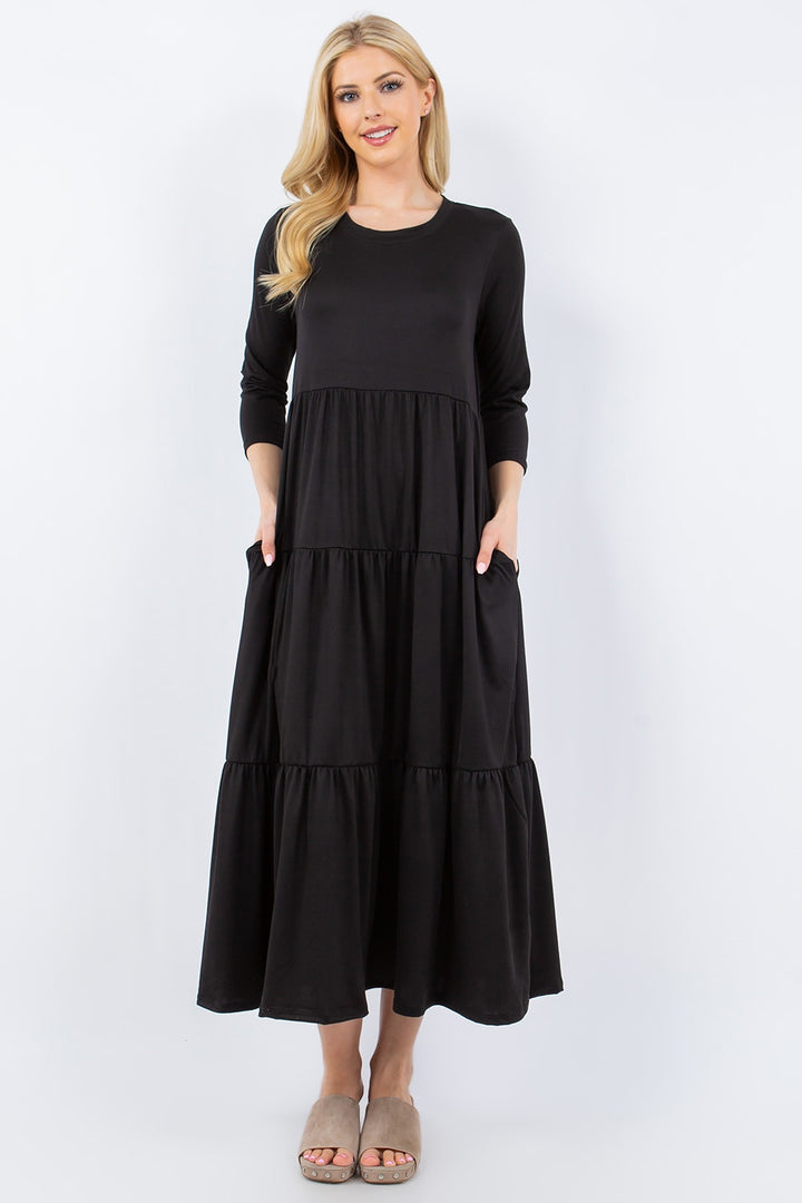 Dressed in the Celeste Full Size Tiered Midi Dress with Pockets, a person stands against a plain white background. This versatile black dress, designed with three-quarter sleeves and a loose fit, pairs perfectly with beige slippers. Its tiered design and handy pockets accentuate the individual's long blonde hair and slight smile.