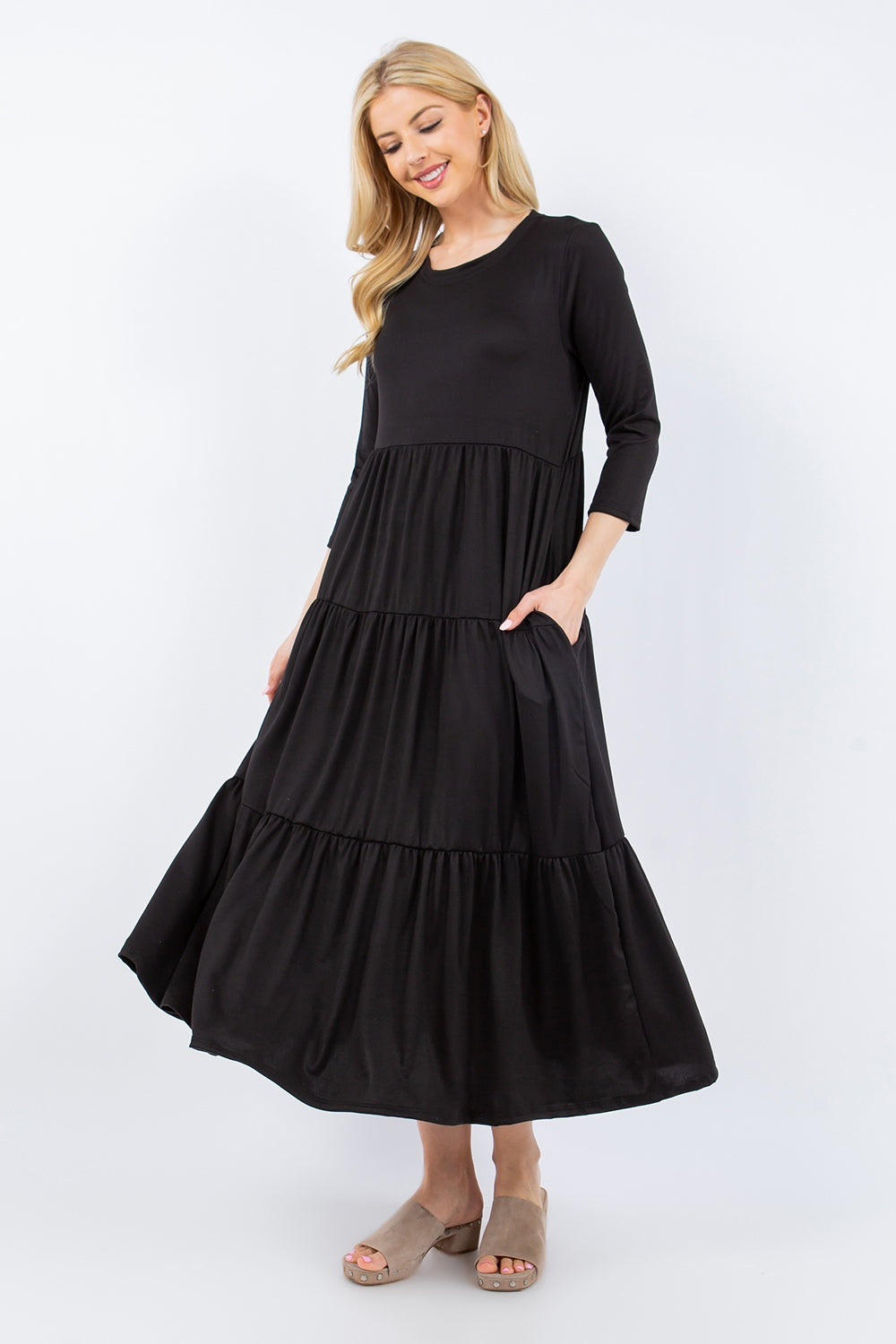 A person poses against a simple backdrop wearing the Celeste Full Size Tiered Midi Dress with Pockets, featuring three-quarter sleeves. Their long hair frames a cheerful expression as they hold the dress to showcase its hidden pockets, while tan slide sandals add an effortlessly chic touch to their outfit.