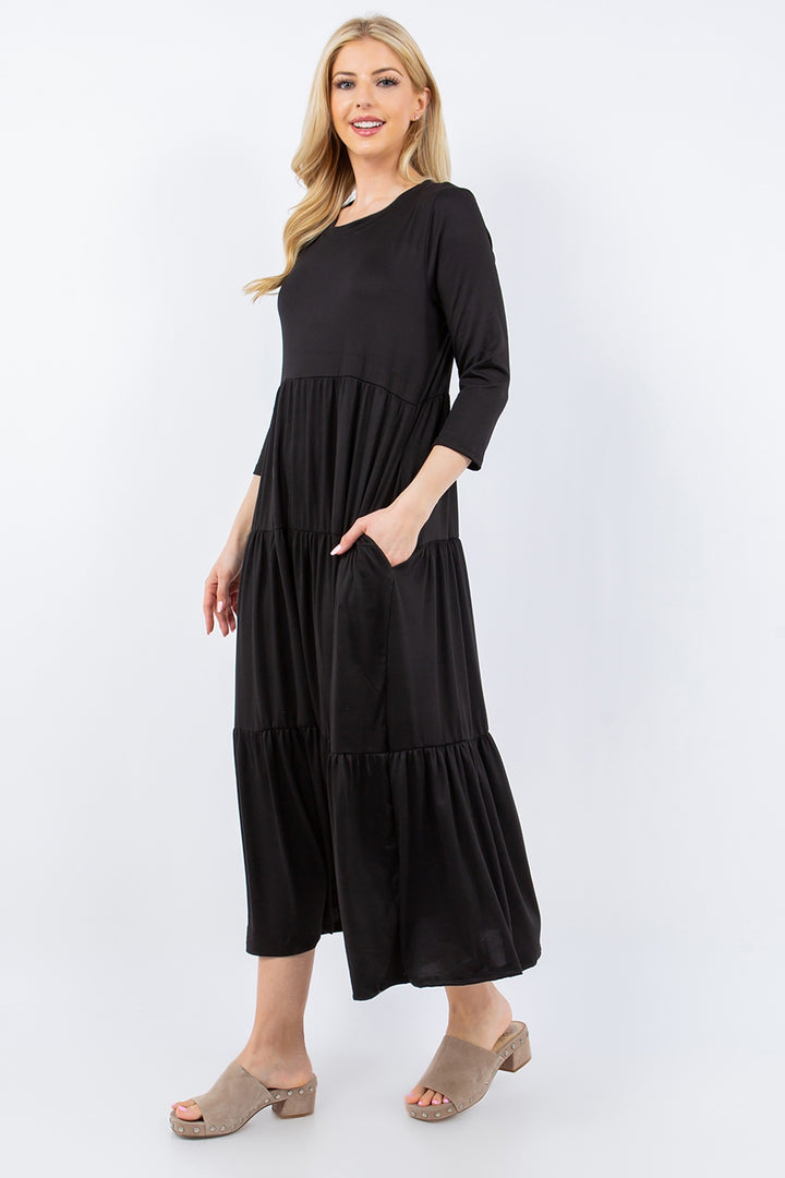 A woman poses in the Celeste Full Size Tiered Midi Dress with Pockets, showcasing its versatile black design and three-quarter sleeves. Her long blonde hair perfectly complements her beige wedge sandals against a plain white background.