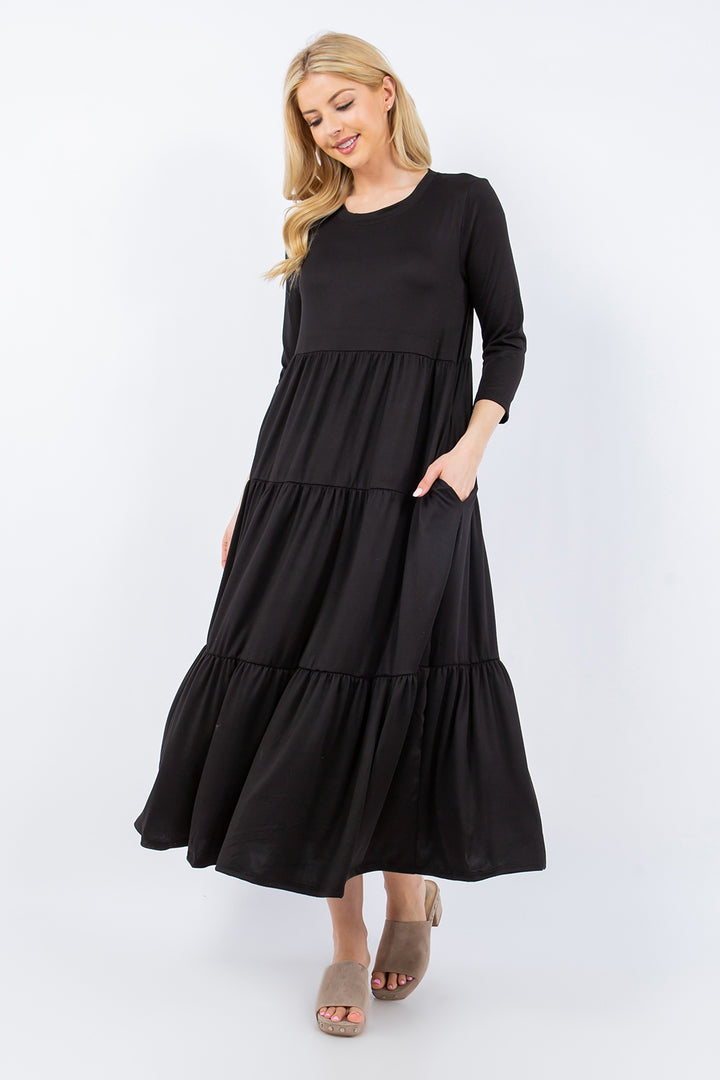 A smiling woman with long blonde hair is wearing the Celeste Full Size Tiered Midi Dress, featuring three-quarter sleeves and convenient pockets, in a versatile black color. She pairs it with beige slip-on shoes while standing against a plain, light background, casually tucking one hand into her pocket.