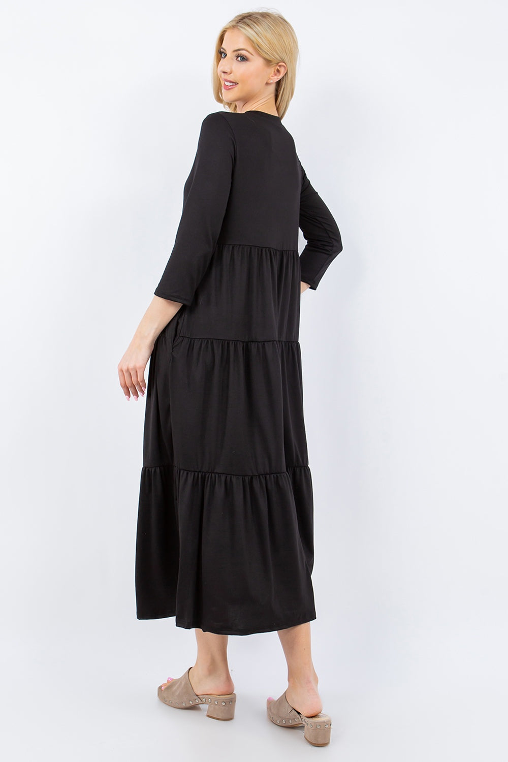 A woman with blonde hair is wearing the Celeste Full Size Tiered Midi Dress with Pockets in black and beige sandals. She stands against a plain white background, facing away from the camera but looking back over her shoulder.