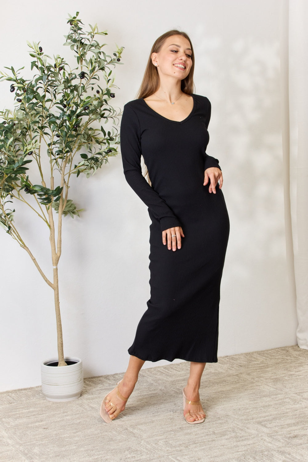 Wearing the Culture Code Full Size Ribbed Long Sleeve Midi Slit Dress, a woman poses gracefully with her right hand on her hip. The chic, form-fitting black dress enhances her style as she stands by a potted plant on a carpeted floor, set against a neutral background.