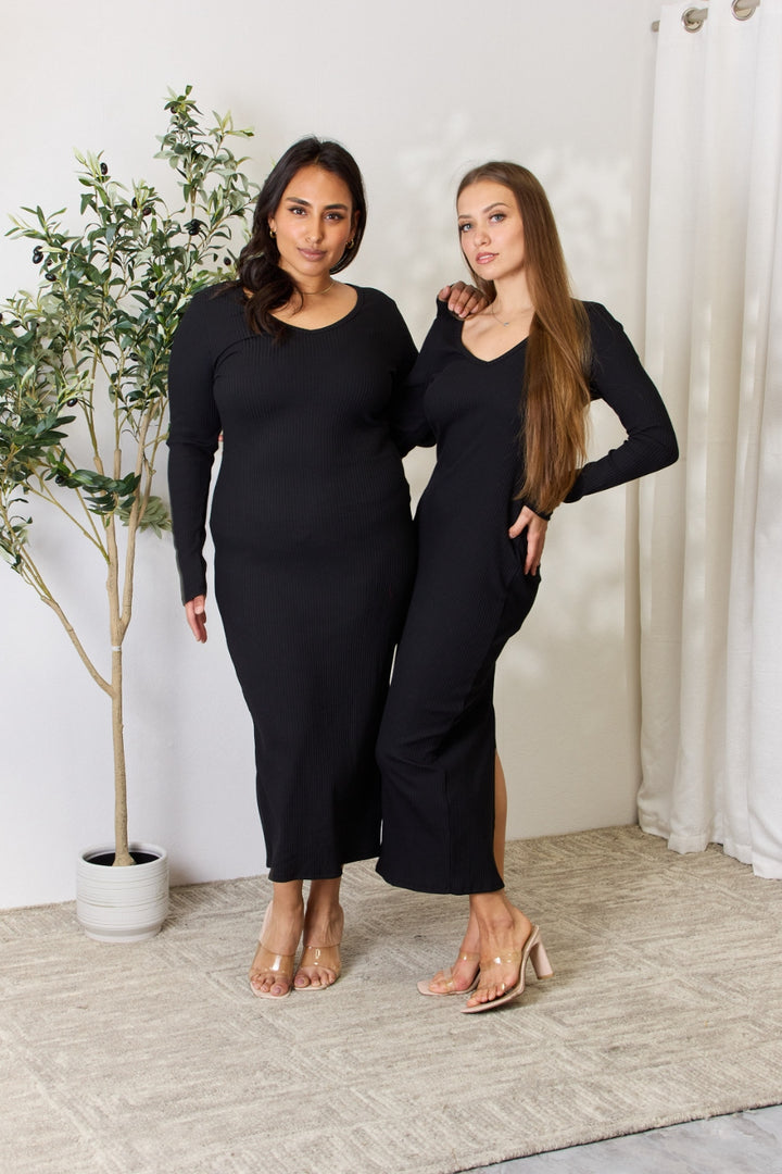 Two women pose confidently side by side in a room, both wearing chic Culture Code Full Size Ribbed Long Sleeve Midi Slit Dresses in black. One has her arm around the others shoulder as they stand on a beige carpet, in front of a small potted plant and white curtain, exuding elegance.