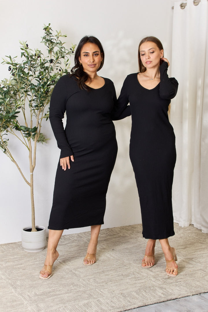 Two women stand side by side in a room with light-colored curtains and a potted plant, both wearing the Culture Code Full Size Ribbed Long Sleeve Midi Slit Dress and sandals. They pose on a neutral-toned woven rug against a plain white wall.