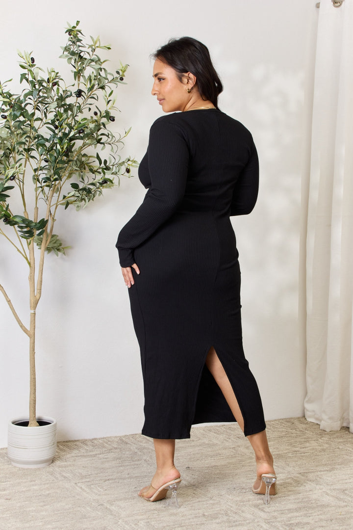 In a well-lit room, someone poses in the Culture Code Full Size Ribbed Long Sleeve Midi Slit Dress, standing by a potted plant on a textured rug with white walls and sheer curtains as the backdrop.