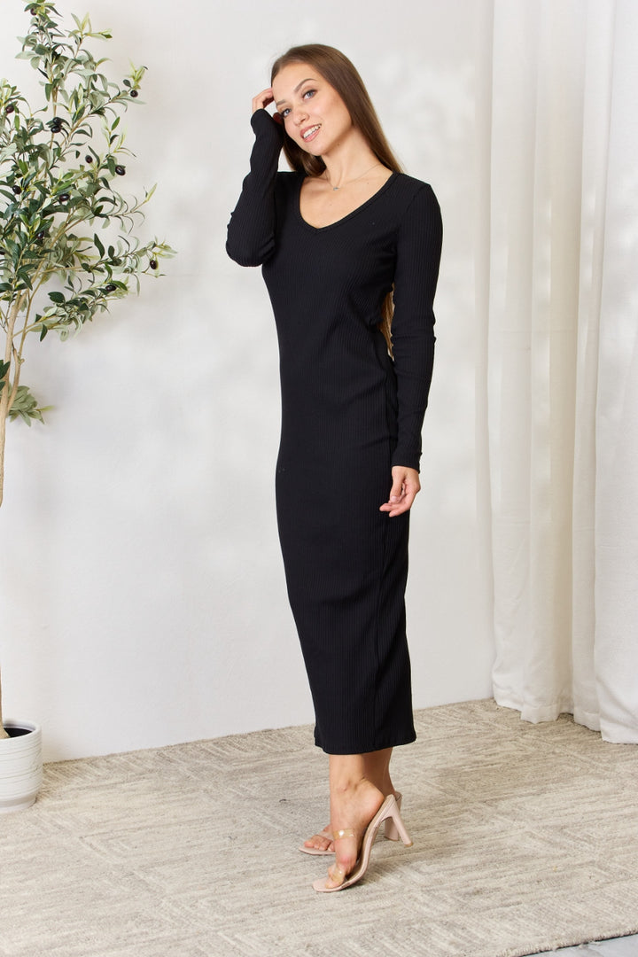 A woman stands on a light carpeted floor in the elegant Culture Code Full Size Ribbed Long Sleeve Midi Slit Dress paired with beige heels. Smiling and touching her hair, she is framed by a potted plant and sheer curtains, adding charm to this versatile fashion moment.