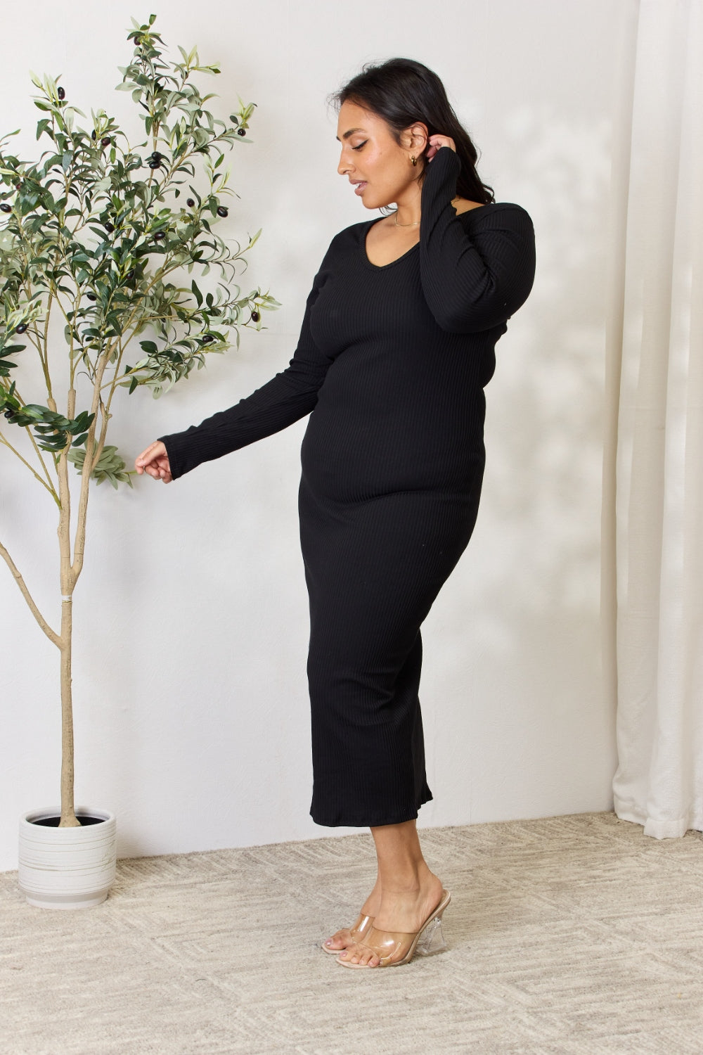 Wearing the Culture Code Full Size Ribbed Long Sleeve Midi Slit Dress and transparent heels, a woman stands by a leafy potted plant. She gazes to the side, touching her hair. The rooms versatile decor includes a light carpet and sheer curtains in the background.