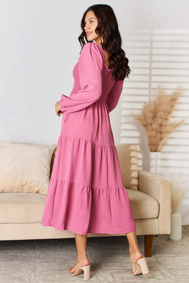 A woman in a Culture Code Full Size Ruffle Trim Smocked Tiered Dress stands in the living room. She is facing sideways and smiling. The room features a beige sofa with cream cushions, and decorative dried pampas grass casting shadows on the wall, adding an elegant touch to the scene.