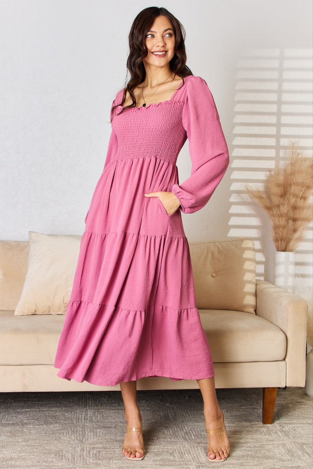 A woman wearing the Culture Code Full Size Ruffle Trim Smocked Tiered Dress, characterized by its long pink fabric, smock detailing, and square neckline, stands in a bright room. The dress features a tiered skirt and long sleeves. She is smiling with one hand in her pocket against the backdrop of a beige couch and decorative pampas grass.