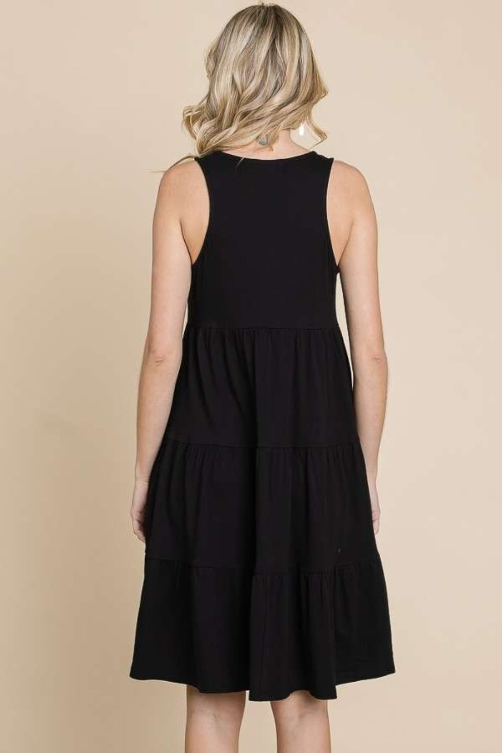 A woman with blonde hair is wearing the Culture Code Racerback Tiered Tank Dress, showcasing its elegant racerback design and tiered details. She is facing away, set against a beige background.