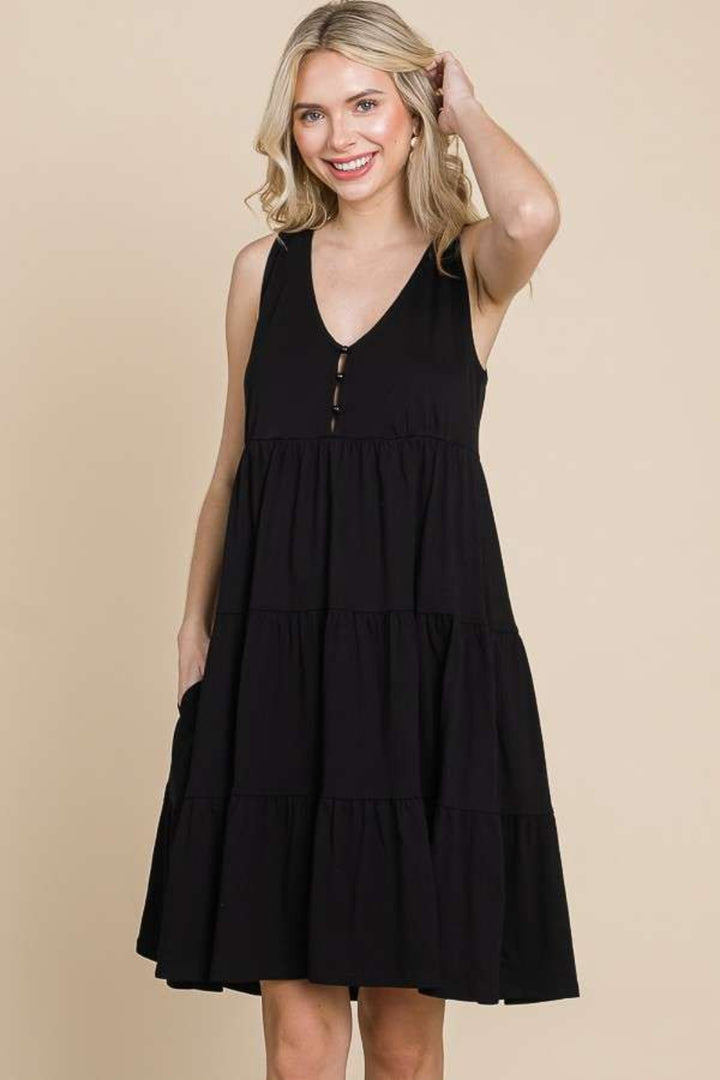 A smiling person with long hair wears a Culture Code Racerback Tiered Tank Dress in black with tiered detailing and front buttons, against a beige backdrop. One hand is on their hip while the other gently touches their hair.