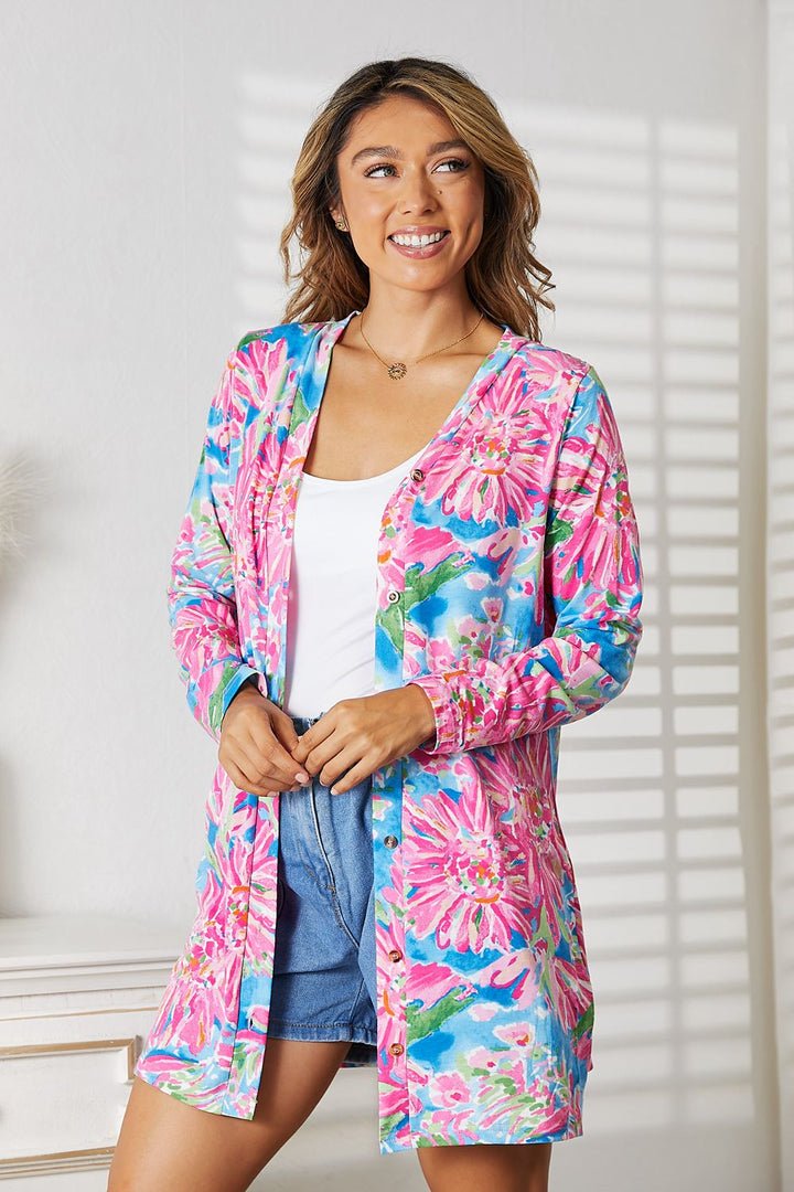 A woman radiates elegance, smiling in a sunlit room. She wears the vibrant Double Take Floral Open Front Long Sleeve Cardigan over a white top and denim shorts. A white dresser and vertical blinds enhance the romantic ambiance with light stripes on the wall.