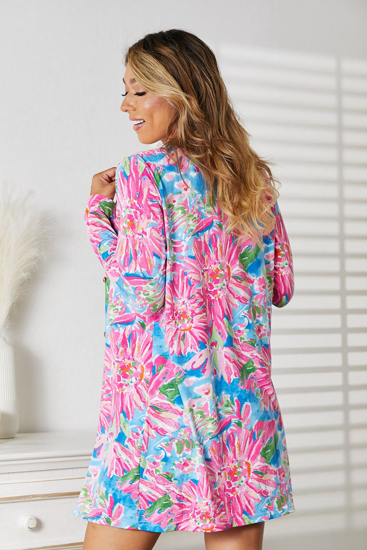 Wearing the Double Take Floral Open Front Long Sleeve Cardigan, a woman with long hair exudes feminine charm. The cardigans vibrant pink, blue, and green floral pattern complements her smile as sunlight casts romantic shadows in the bright room where she stands with her back slightly turned.