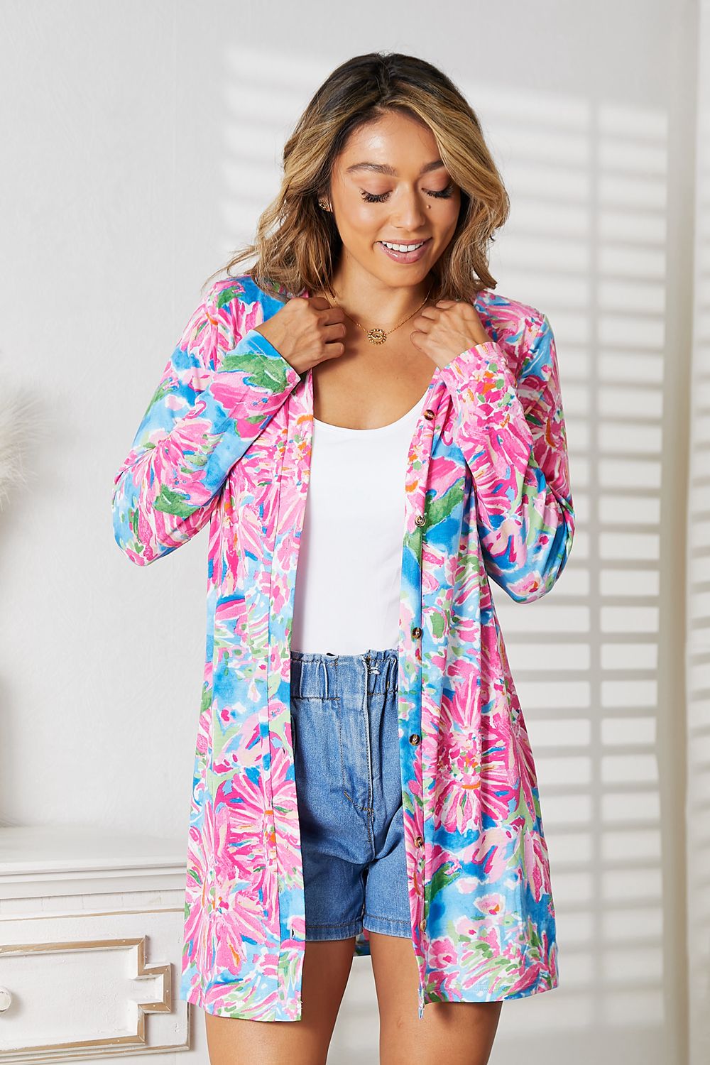 A woman smiles, radiating charm in the Double Take Floral Open Front Long Sleeve Cardigan over a white top and denim shorts. She gently adjusts it, while the soft shadows from a window on a white wall enhance her romantic elegance.