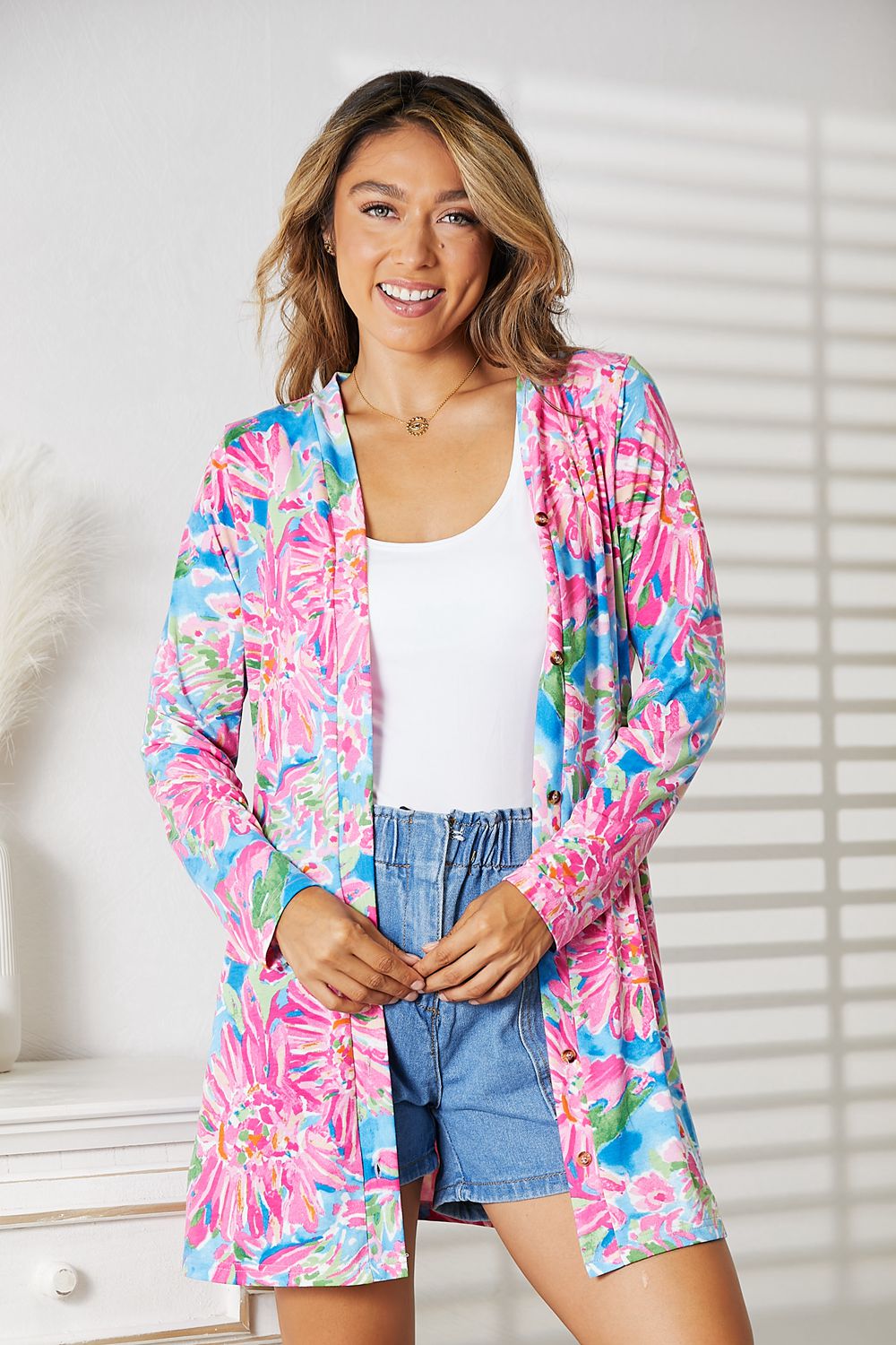 A person wears the Double Take Floral Open Front Long Sleeve Cardigan over a white top and denim shorts indoors, radiating feminine charm. Soft natural light and shadow patterns from a nearby window enhance the romantic, elegant look with their gentle interplay.