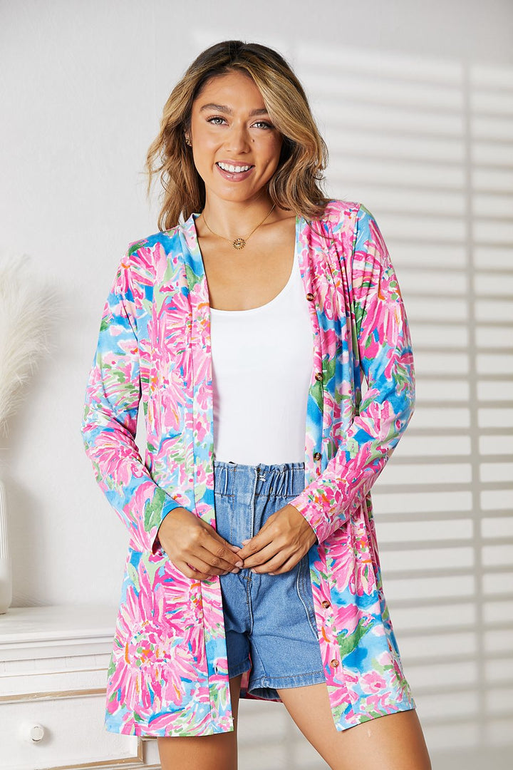 A person wears the Double Take Floral Open Front Long Sleeve Cardigan over a white top and denim shorts indoors, radiating feminine charm. Soft natural light and shadow patterns from a nearby window enhance the romantic, elegant look with their gentle interplay.