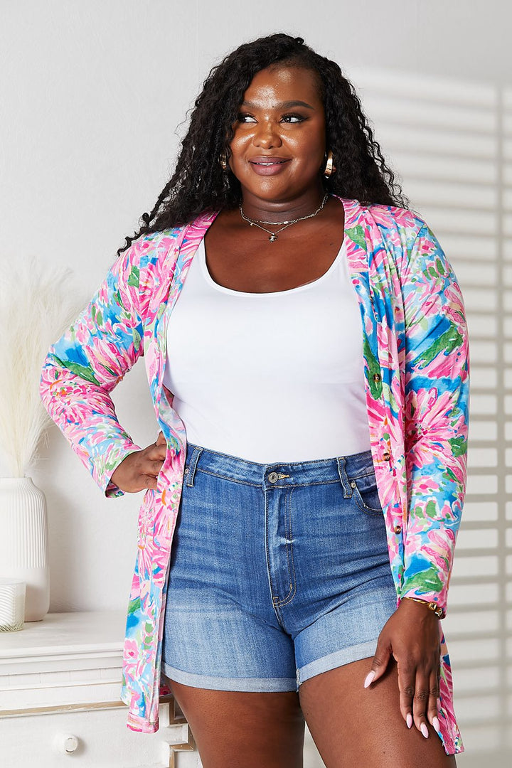 A woman exudes charm in a Double Take Floral Open Front Long Sleeve Cardigan over a white top and denim shorts. Her long curly hair enhances her romantic look in a bright room, highlighted by decorative touches like a vase on the cabinet.