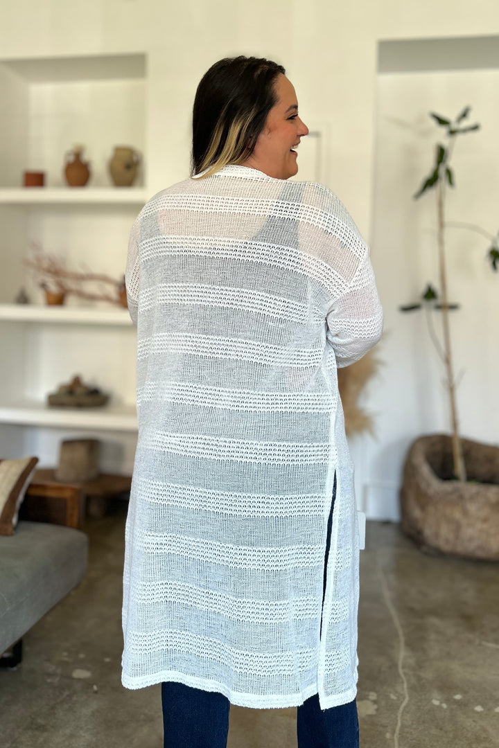 A person is standing indoors, facing away from the camera, wearing a long Double Take Full Size Open Front Longline Cardigan over blue jeans. The room features a gray couch, white shelves adorned with decorative items, and a plant in the background.