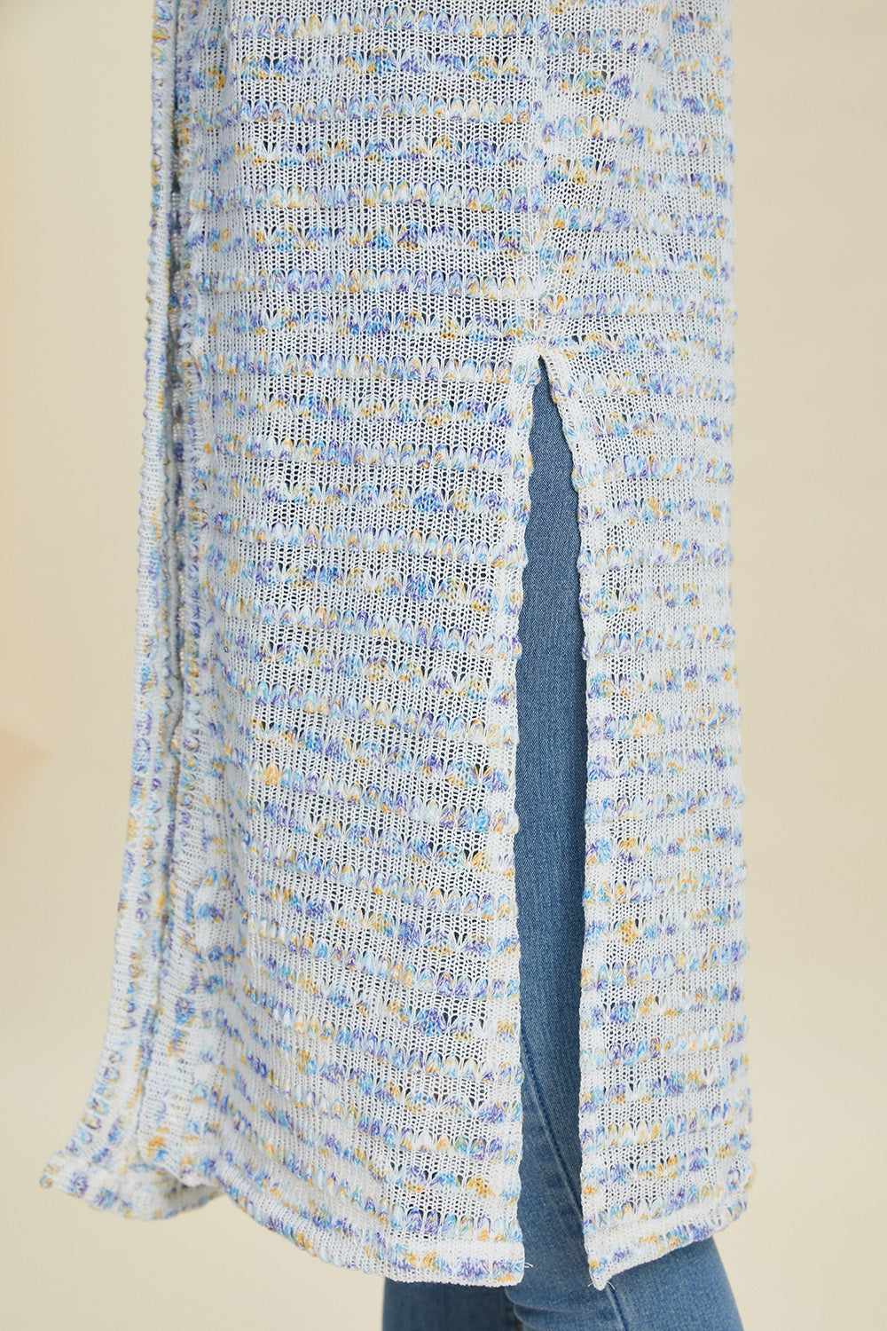 Close-up of the Double Take Full Size Open Front Longline Cardigan, featuring a slit that reveals denim beneath. The cardigan showcases an openwork textured pattern in blue, white, and hints of orange set against a neutral background. It subtly incorporates stretch elements for added comfort and fit.