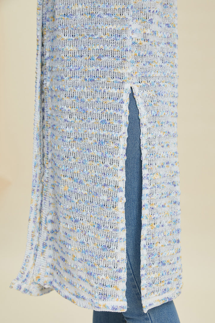 Close-up of the Double Take Full Size Open Front Longline Cardigan, featuring a slit that reveals denim beneath. The cardigan showcases an openwork textured pattern in blue, white, and hints of orange set against a neutral background. It subtly incorporates stretch elements for added comfort and fit.