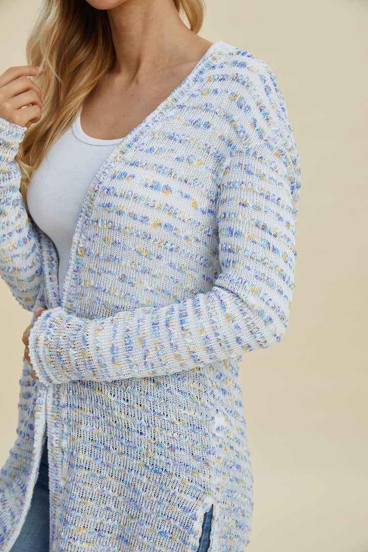 A woman wearing the Double Take Full Size Open Front Longline Cardigan, made of soft polyester with a textured, multicolored design, drapes gracefully over a white top. Featuring a stylish blend of light blue, yellow, and white hues, this piece is perfect for any season. Her long blonde hair cascades as her hand gently rests on the cardigan.