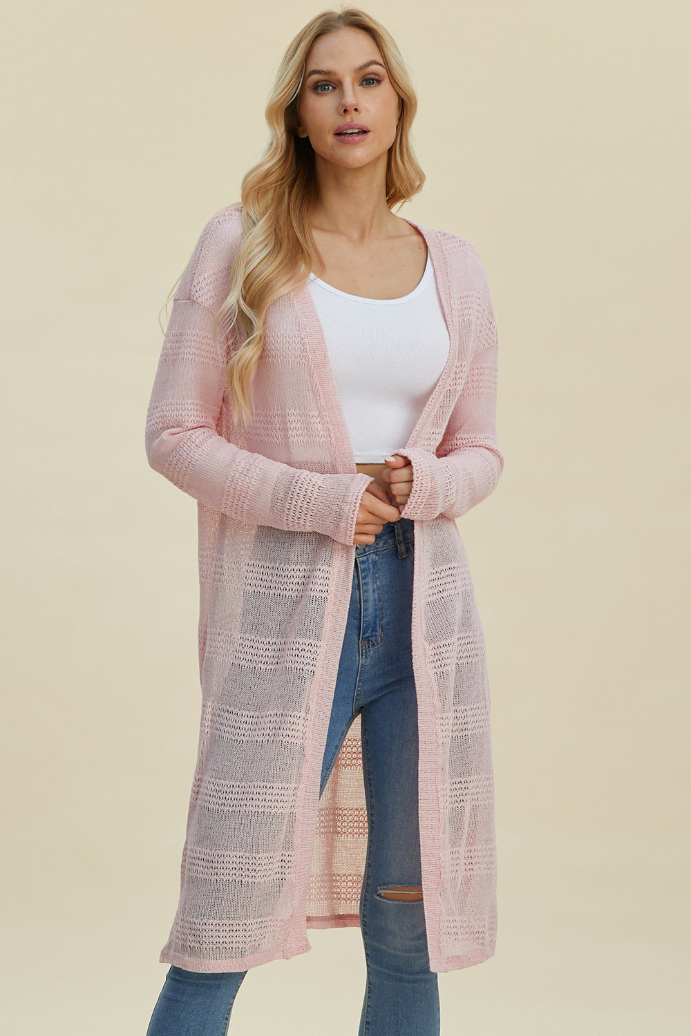 A person with long blonde hair is wearing a white top, blue jeans, and the Double Take Full Size Open Front Longline Cardigan in light pink. Standing against a beige background, the knee-length cardigan is open to reveal its knitted texture and comfortable hint of stretch.